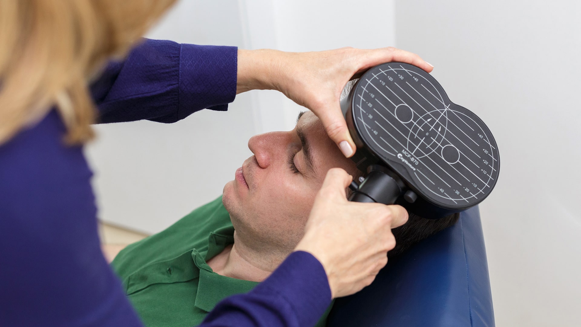 Mãos de uma pessoa segurando um aparelho de formato oval com um cabo, próximo à cabeça de um homem com expressão serena